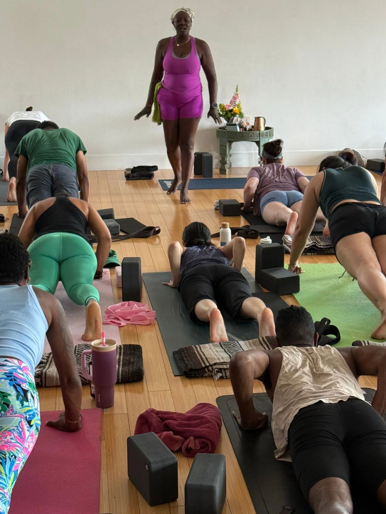 A yoga instructor leads a diverse class through a plank pose in a bright studio.