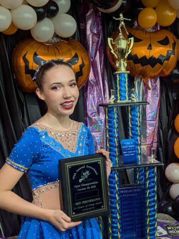 Young girl in blue dance costume holding a plaque and large trophy at a competition.