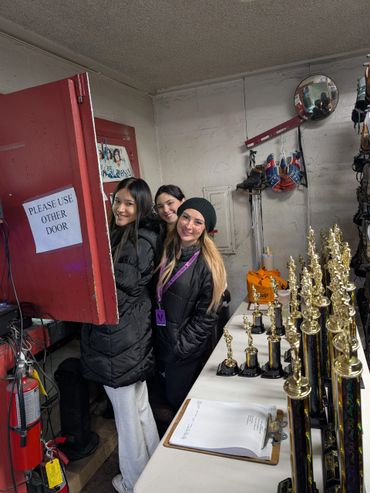 Three women posing behind a door near a table with trophies and a clipboard.