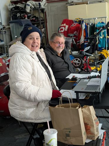 Two people sitting at a table with a laptop and grocery bags in a garage filled with sports gear.