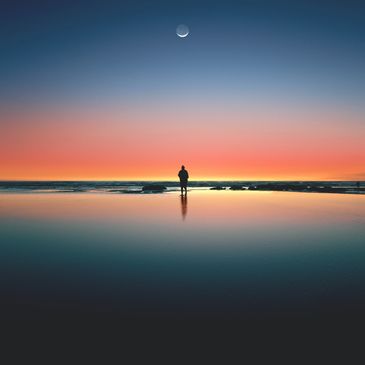 A lone person stands by the ocean at sunset under a crescent moon