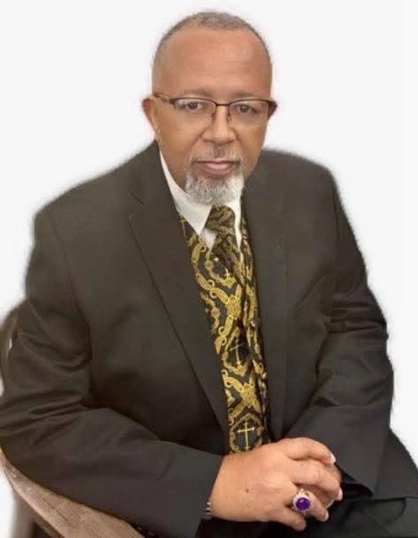 A distinguished man in a suit and patterned tie sitting with hands clasped.