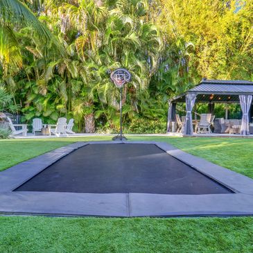 In-ground trampoline with basketball hoop in a lush backyard.