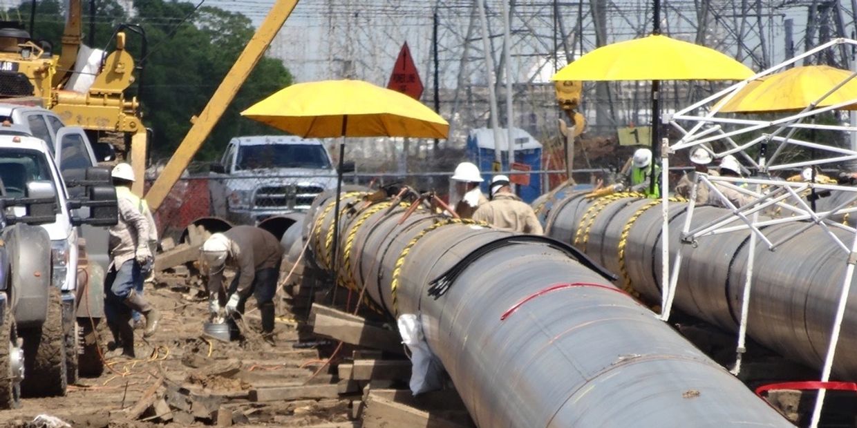 Construction workers install large industrial pipelines under yellow umbrellas.