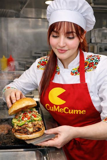 Chef in a red apron holding a large gourmet burger with fresh toppings.