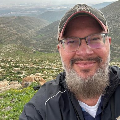 Smiling man with glasses and cap in a mountainous area.