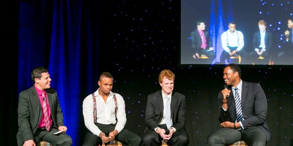 Four men in suits engage in a lively panel discussion on stage.
