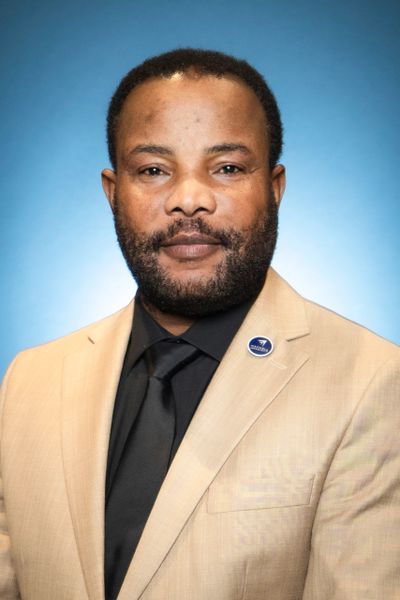 Professional portrait of a man in a beige suit and black shirt with a blue background.
