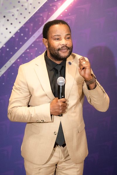 Man in beige suit speaking into a microphone on stage.