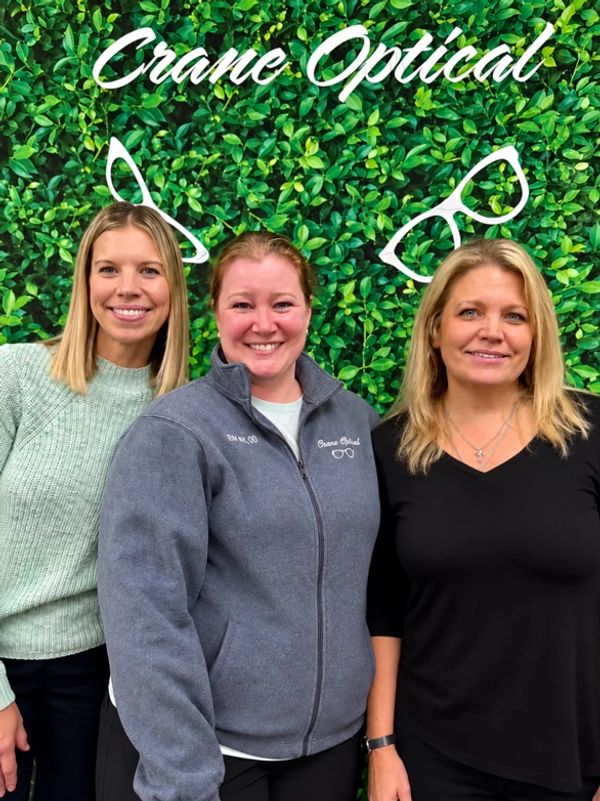 Three women smiling in front of a green leafy wall with 'Crane Optical' sign.