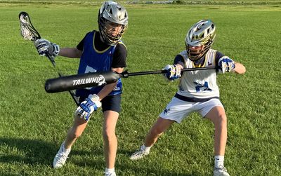 Two young lacrosse players wearing helmets and protective gear practicing with a Tailwhip training stick on grass.