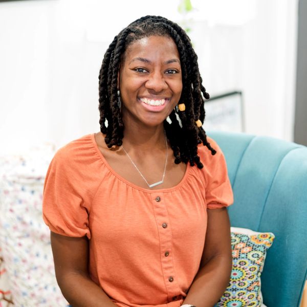 Smiling woman with braided hair wearing an orange top, sitting on a blue chair.