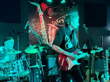 A guitarist and drummer perform on stage with musical decor in the background.