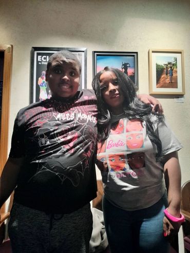 A young man and woman posing together indoors with framed pictures behind them.