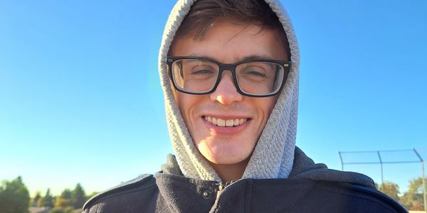 Young man smiling in sunlight with glasses and hoodie.