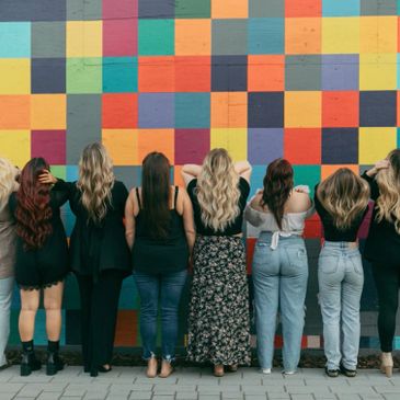 Seven women standing back to colorful checkered wall, showing diverse hairstyles.