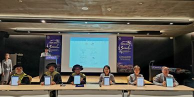 Six people seated at a table holding up tablets in front of a presentation screen.