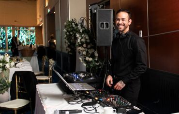 A smiling DJ stands behind his mixing equipment at an elegant event venue.