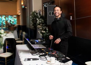 A smiling DJ stands behind a mixing console at an event venue.