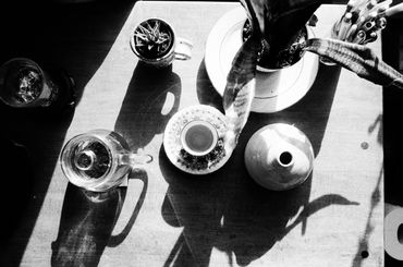 Black and white overhead still life of items on desk, including glass teapot, plants and teacup