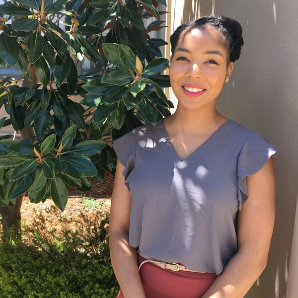 Young woman smiling outdoors near a leafy plant on a sunny day.