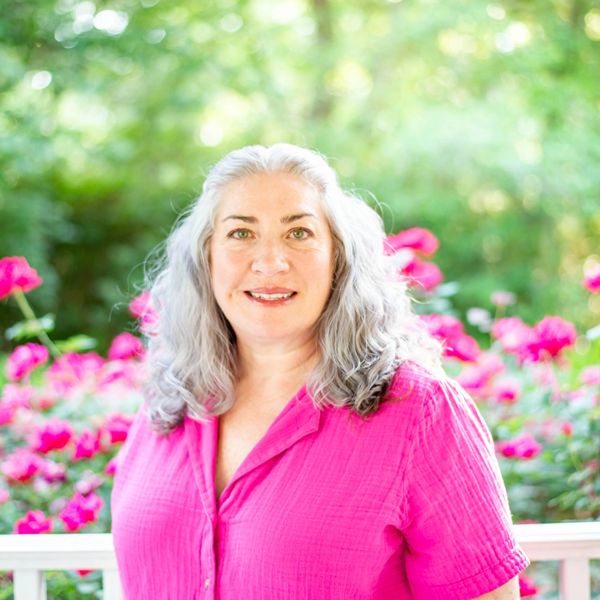 Woman with gray hair in a bright pink shirt smiles outdoors among pink flowers.