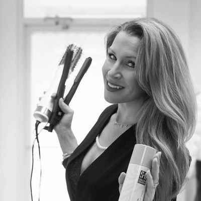 Woman holding hair styling tools and product, smiling.