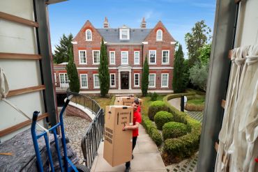 A person moving large boxes outside a grand brick house.