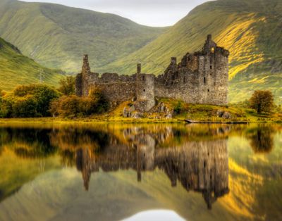 Ancient castle ruins reflected in a calm lake surrounded by green hills.