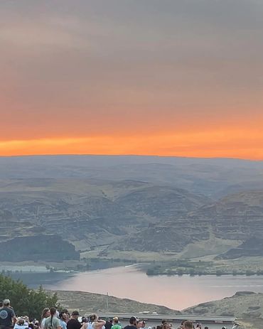 Crowd gathered near a river with hills and a colorful sunset in the background.