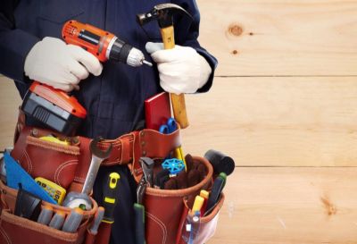 Person in work gloves holding a drill and hammer with a tool belt full of tools.