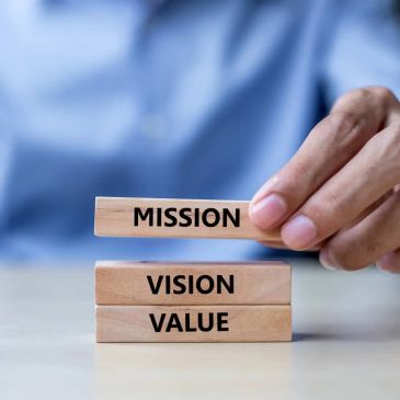 Hand stacking wooden blocks labeled Mission, Vision, Value.