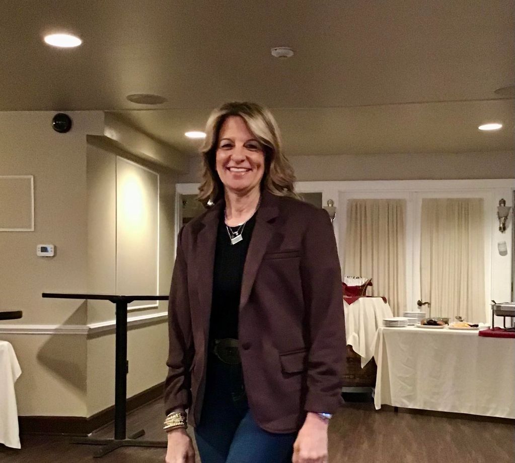 Smiling woman in a dark blazer standing in a warmly lit room.