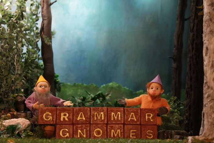 whimsical gnomes in a forest behind a brick wall. Letters on the wall spell the words Grammar Gnomes