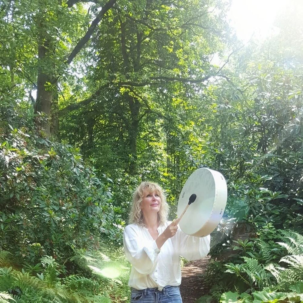 Woman playing a drum in a sunlit forest surrounded by greenery.