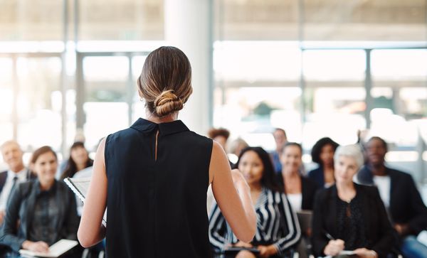 A woman with a bun addresses an audience from the back, in a well-lit room