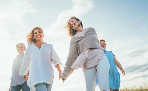 Four joyful women walking together under a bright sky, two holding hands