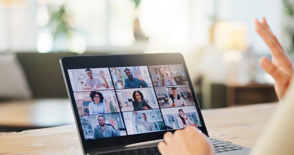 A person's back to the camera, waving at a laptop screen displaying a video conference
