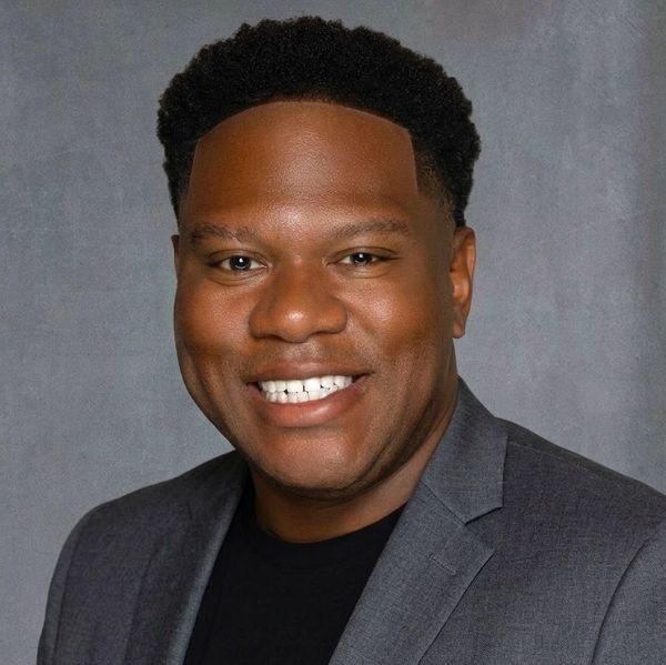 Smiling man in a gray suit jacket and black shirt against a gray background.