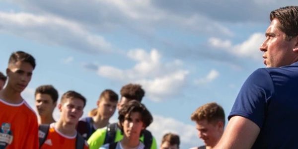 Coach addressing a group of young soccer players outdoors under a partly cloudy sky.