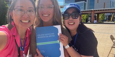 Three women smiling outdoors, holding a "College Success and Advanced ESL Writing" booklet.