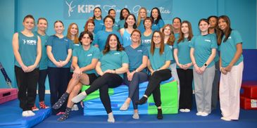 Group photo of young gymnasts and coaches in blue KiPS T-shirts at a gymnastics center.