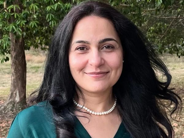 Smiling woman with long black hair wearing a green top and pearl necklace outdoors.