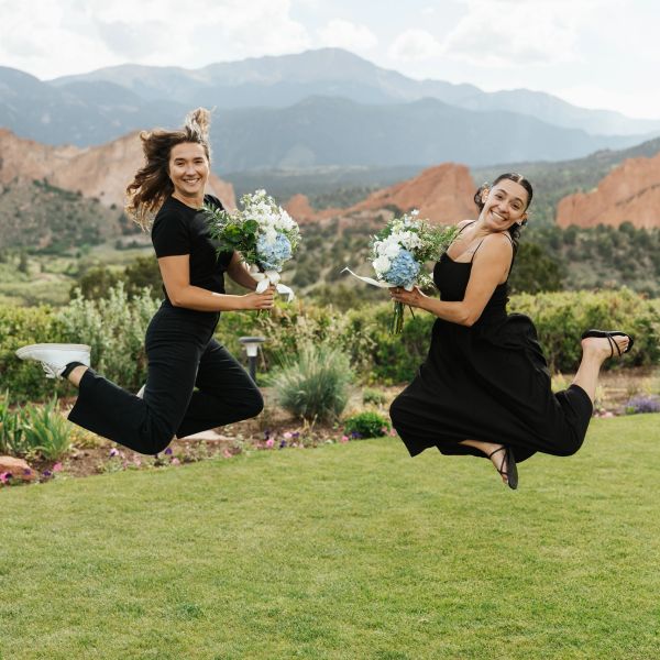 Two happy women jumping with bouquets in a scenic garden.