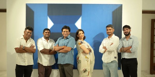 Six people standing confidently with arms crossed in front of a blue geometric painting.