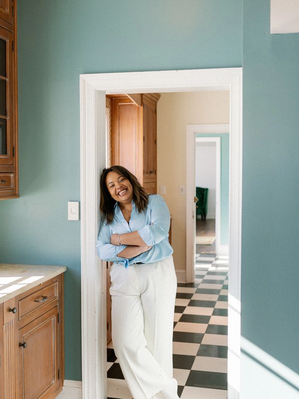 Smiling woman in light blue shirt and white pants leaning in doorway.