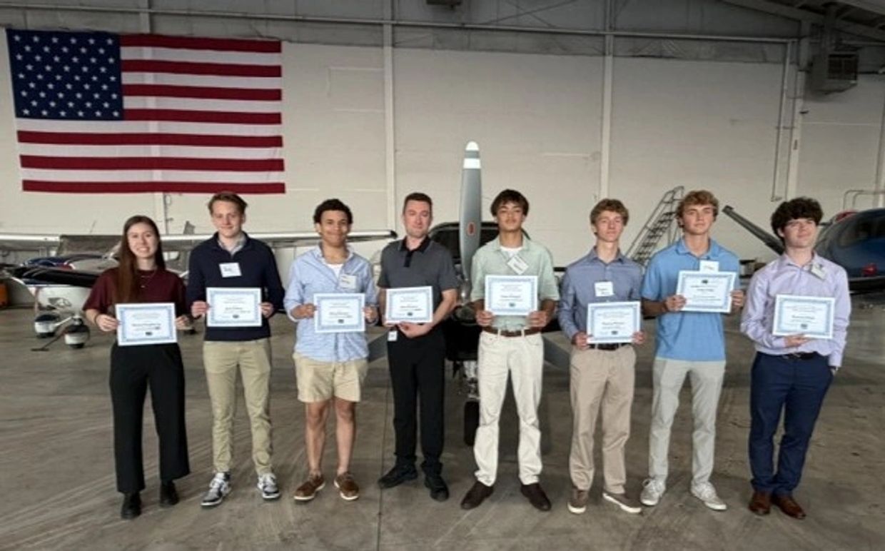 Group of young people holding certificates in an aircraft hangar under an American flag.