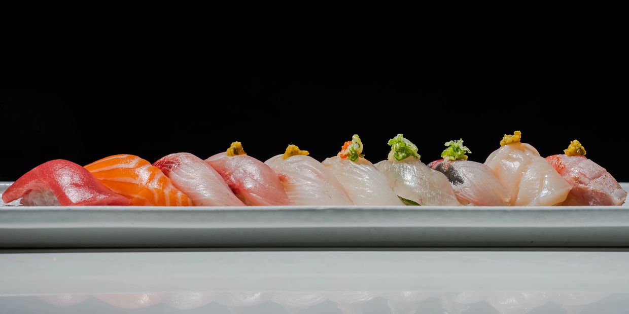 A row of assorted nigiri sushi on a white plate against a black background.