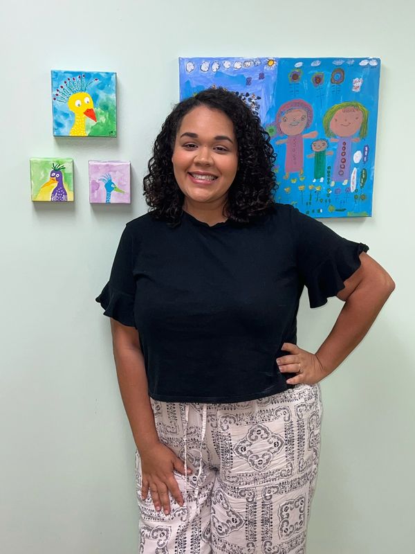 Smiling woman posing in front of colorful childlike paintings on a white wall.