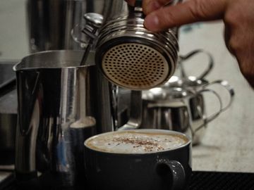 A freshly made cappucino coffee being dusted with chocolate powder by the barista before serving.  In the background are a milk jug of hot frothed milk and a milk thermometer. 